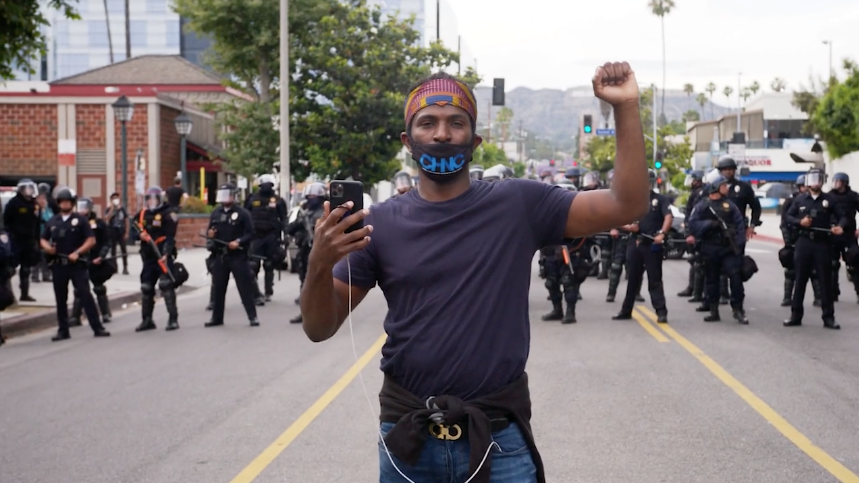 Video screenshot of man, staring into the camera with a fist raised, as he stands in front of a line of police officers.